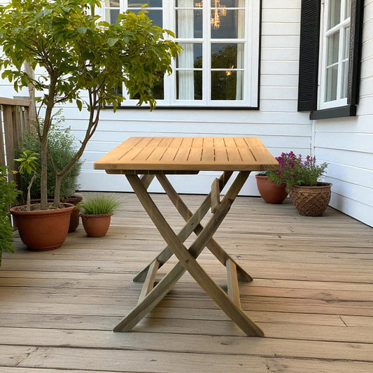 Cilacap square folding table in teak wood, placed on a sunlit patio with potted plants and a white house backdrop featuring black shutters and a large window