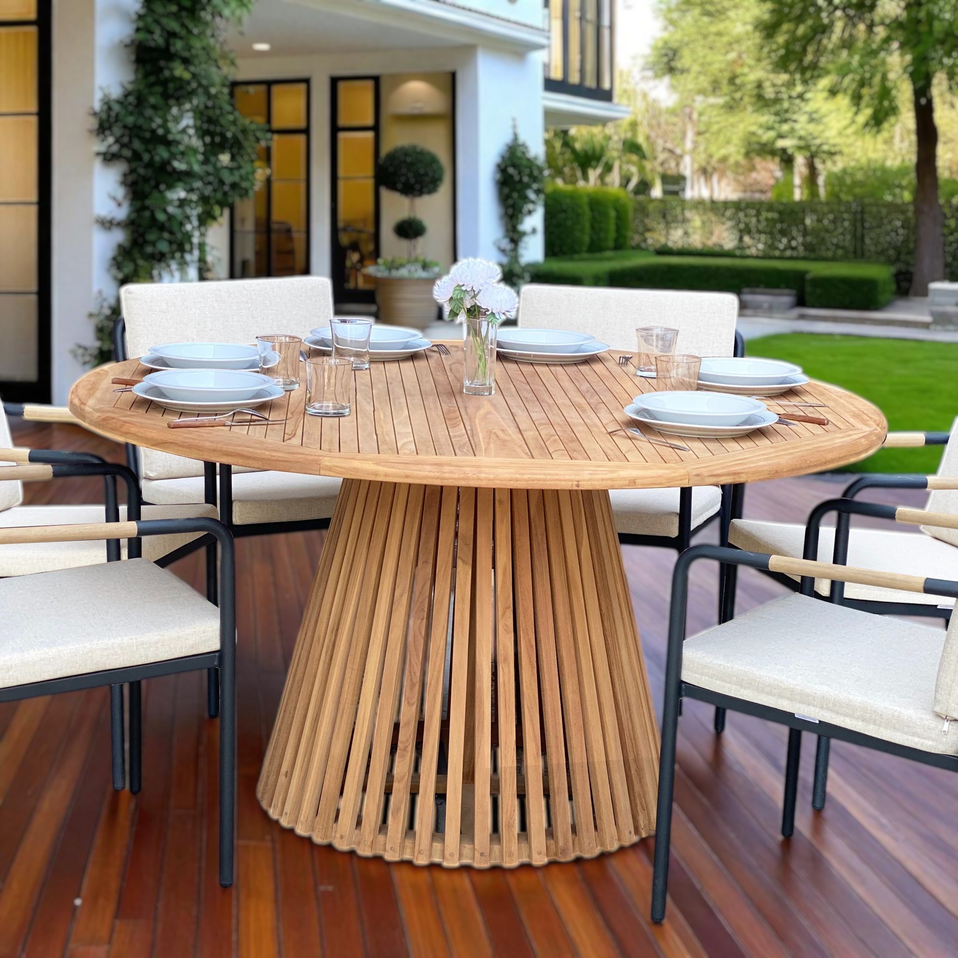 Nusa Dua round wooden outdoor dining table with slatted base, styled with white tableware and floral centerpiece on a garden patio deck.