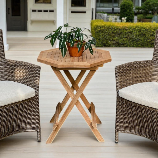 Teak wood Cilacap Side Table with octagonal top, styled between two wicker chairs on a wooden patio, surrounded by lush potted plants and glass-panelled doors—ideal for outdoor relaxation.