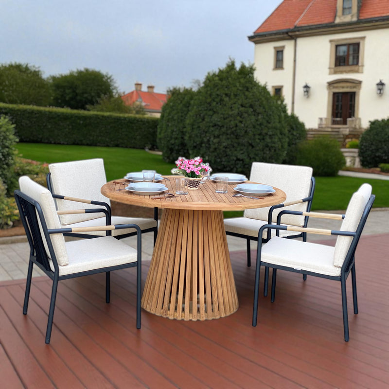 Nusa Dua round wooden outdoor dining table with slatted base paired with Hollviken chairs featuring black metal frames and beige cushions, styled on a wooden patio deck surrounded by greenery—perfect for modern outdoor dining.