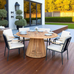 Nusa Dua round wooden outdoor dining table with slatted base paired with Hollviken chairs featuring black metal frames and beige cushions, styled on a wooden patio deck surrounded by greenery—perfect for modern outdoor dining.