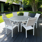 Pisa Outdoor Dining Set featuring a round grey slatted table with white aluminum X-base and six matching chairs, placed on a dark wood patio surrounded by greenery and a modern glass-fronted building.
