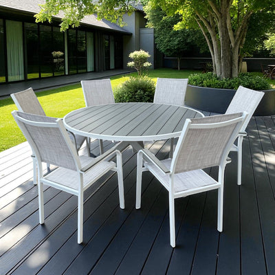 Pisa Outdoor Dining Set featuring a round grey slatted table with white aluminum X-base and six matching chairs, placed on a dark wood patio surrounded by greenery and a modern glass-fronted building.