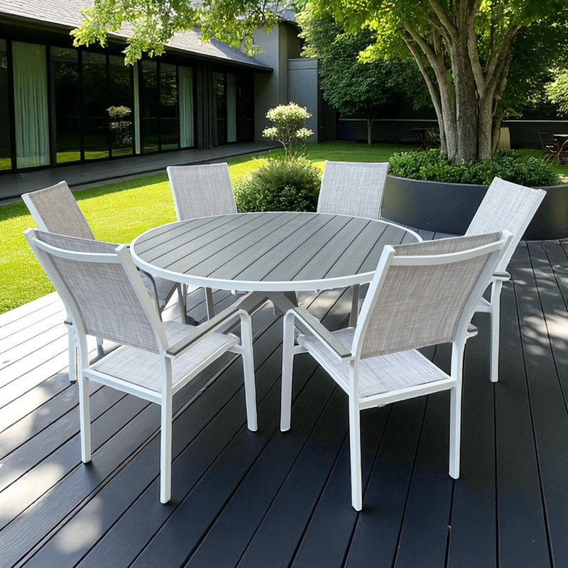 Pisa Outdoor Dining Set featuring a round grey slatted table with white aluminum X-base and six matching chairs, placed on a dark wood patio surrounded by greenery and a modern glass-fronted building.